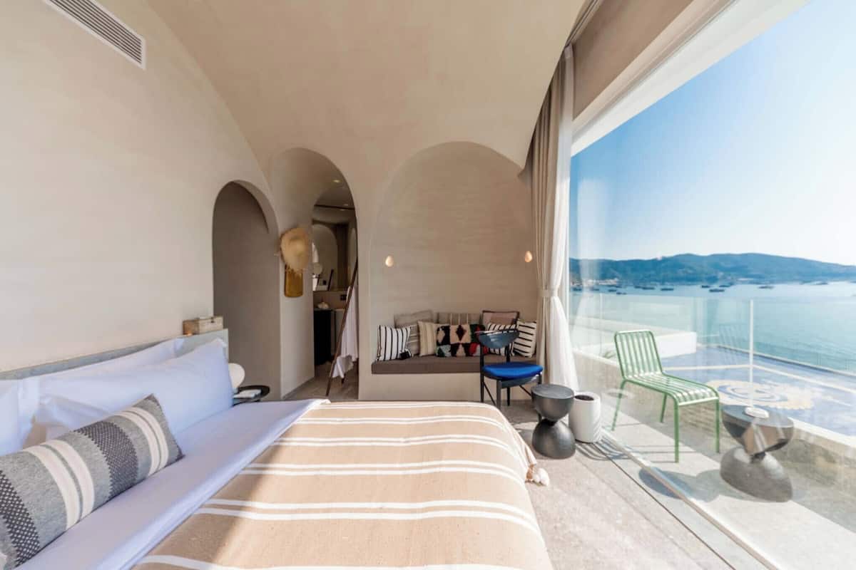 Une chambre d’hôtel moderne dotée d’un grand lit, d’un coin salon avec vue sur la mer et d’un balcon avec une balustrade en verre.