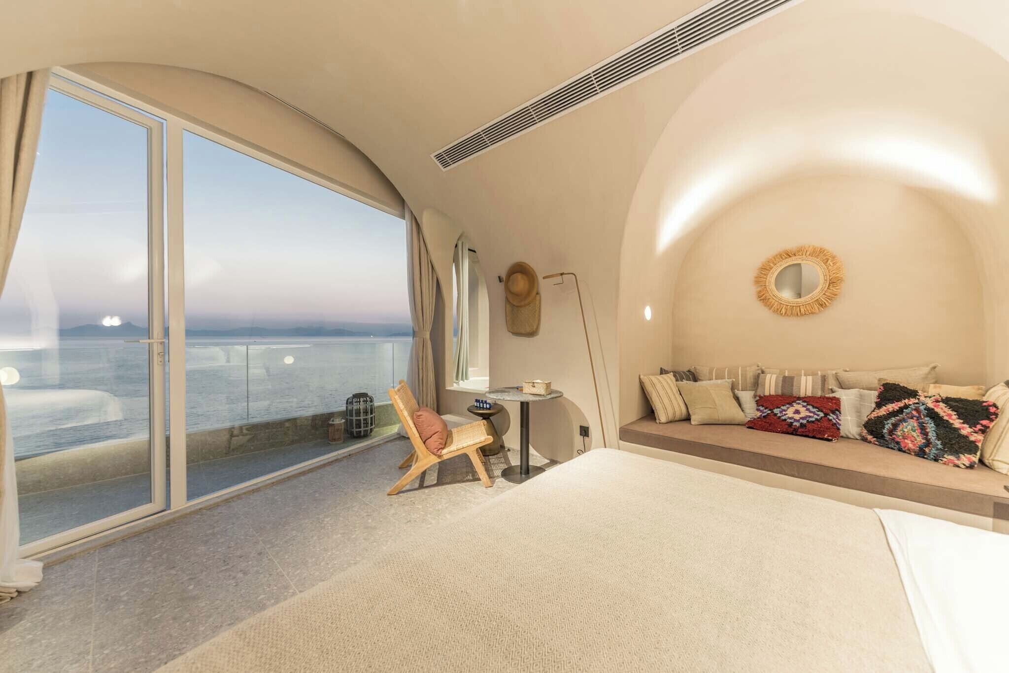 A modern bedroom with a large window, a round mirror, a sofa with colorful cushions, and a small table.