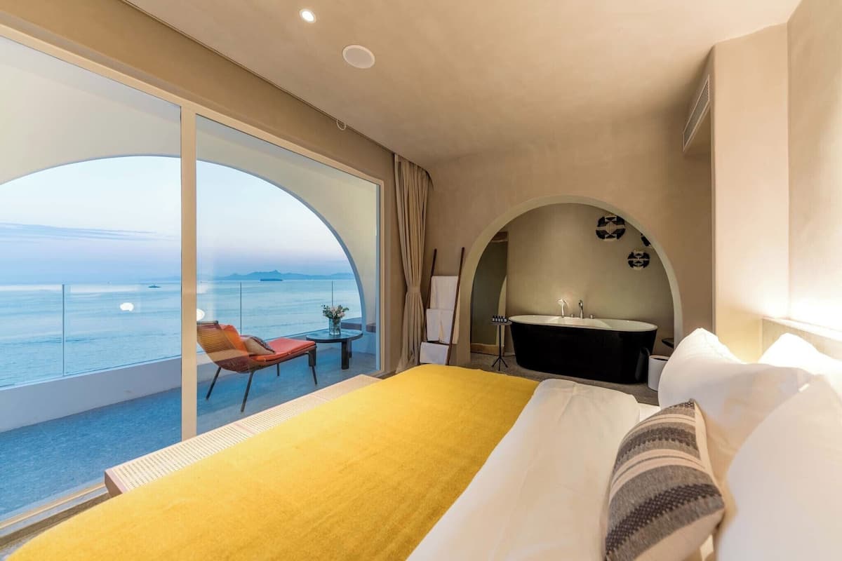 Une chambre d’hôtel avec un grand lit, une vue sur la mer, une salle de bain avec baignoire et une petite table avec une chaise.