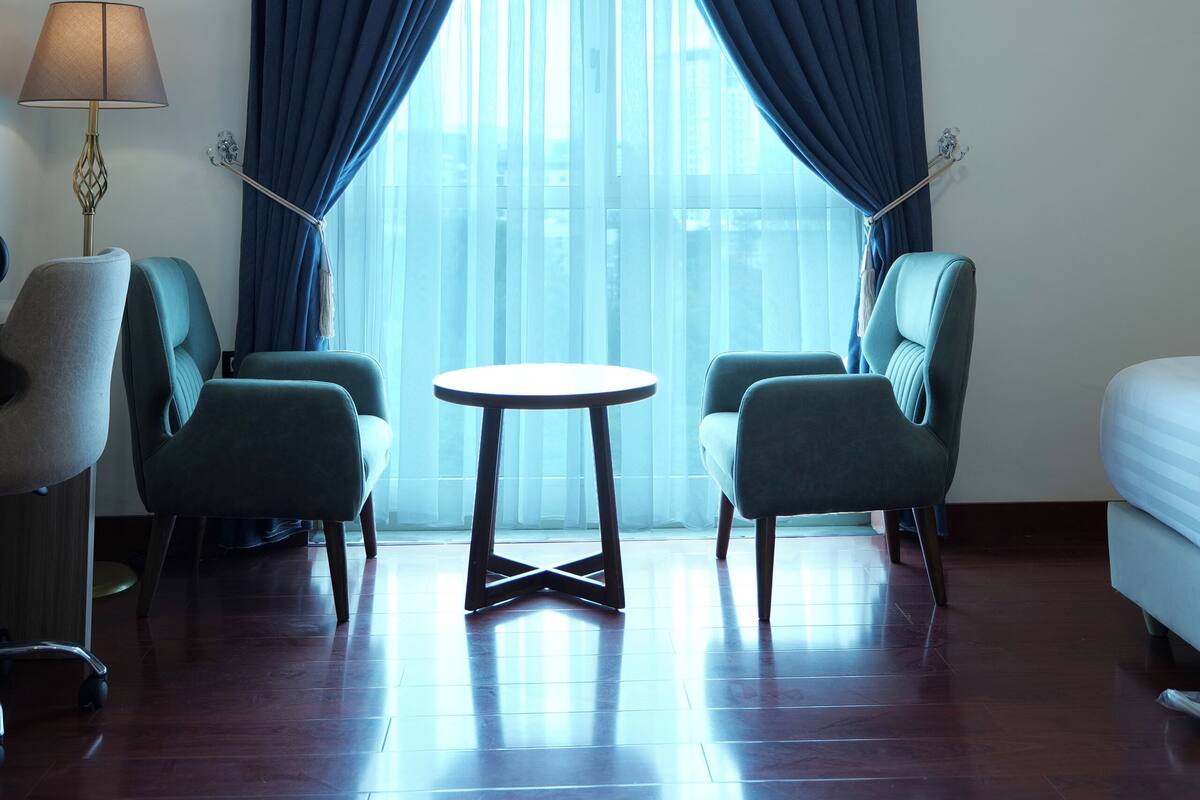 A hotel room with a large window, two chairs, a small table, and a lamp.