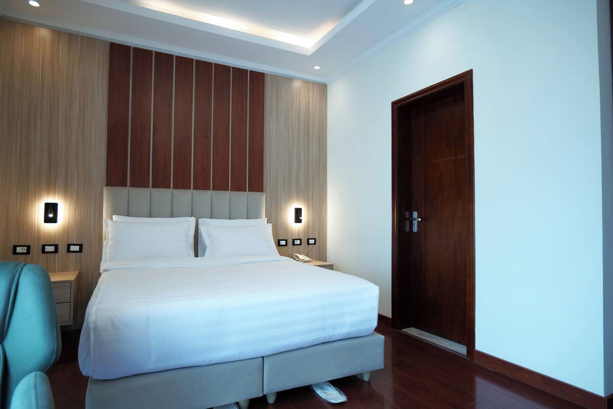 A modern hotel room with a large bed, wooden panel wall, two bedside lamps, and a wooden door.