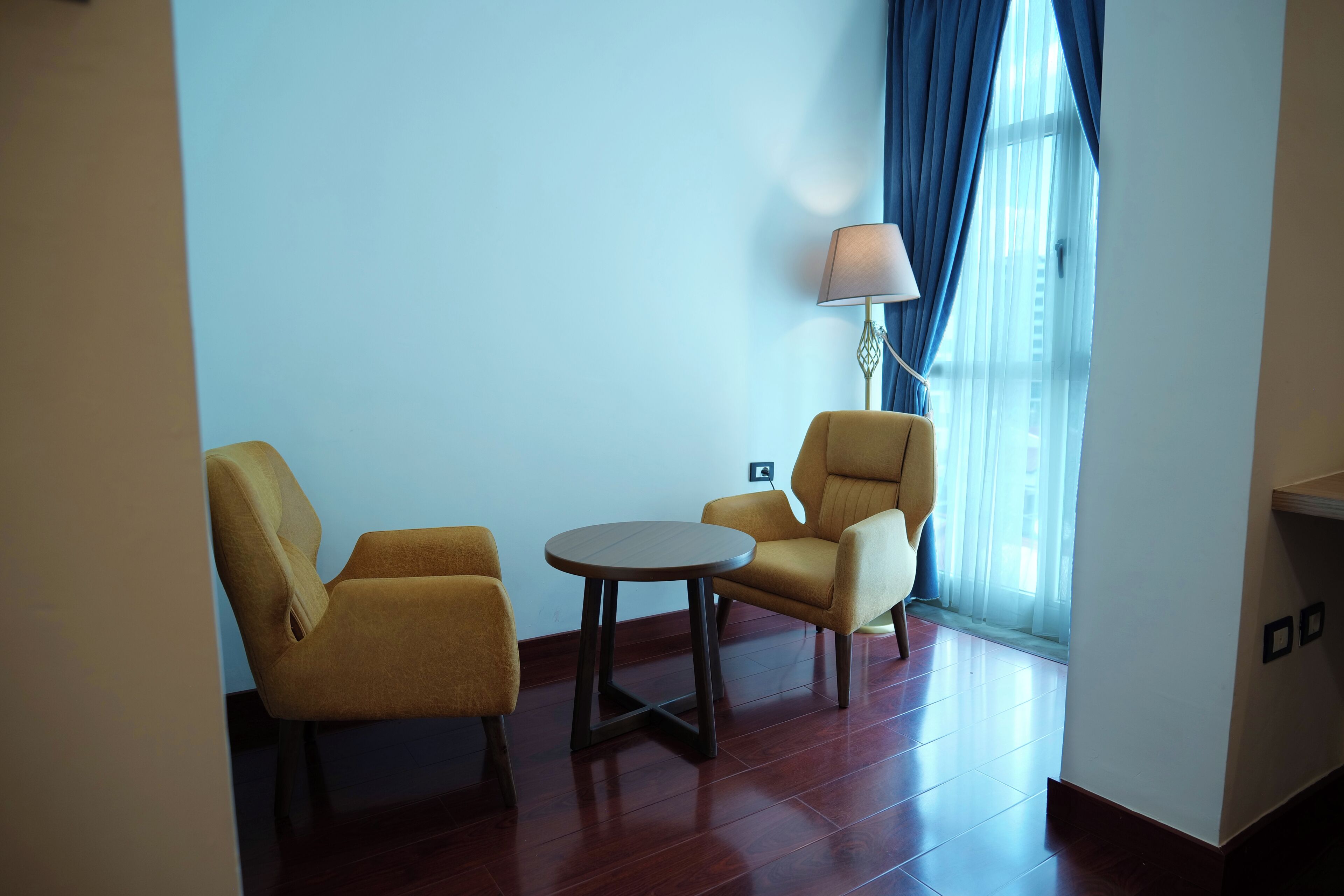 A room with two yellow armchairs, a small round table, a lamp, and a window with blue curtains.