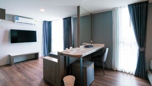 Habitación de hotel moderna con piso de madera, un escritorio con silla, un refrigerador pequeño, un cesto de basura, un televisor de pantalla plana montado en la pared y una ventana con cortinas.