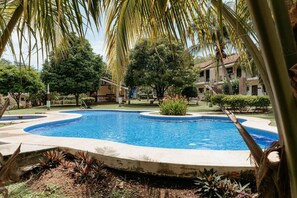 A swimming pool with a curved edge, surrounded by palm trees and greenery, with a building in the background.