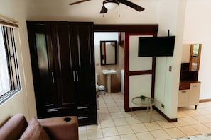 Ein Hotelzimmer mit Schrank, einem kleinen Tisch mit Pflanze, einem an der Wand montierten Fernseher, einem Badezimmer mit Waschbecken und Toilette sowie einem Deckenventilator.