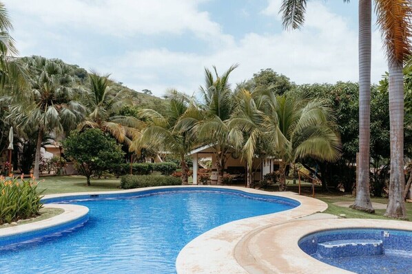 Ein Pool mit geschwungener Kante, umgeben von Palmen und Grünpflanzen, mit einem Gebäude im Hintergrund.