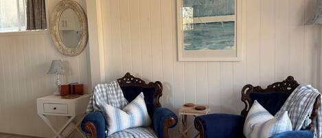 A room with two blue armchairs, a white table, a small side table, a mirror, and a painting on the wall.