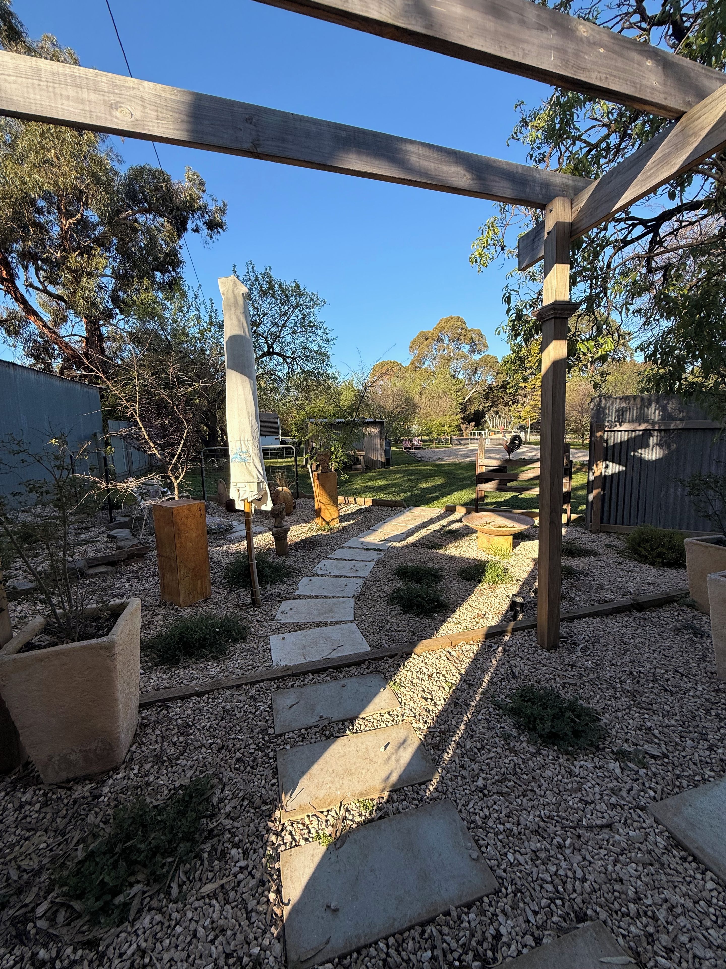 Un giardino con un vialetto in pietra, una pergola in legno e diverse piante in vaso.