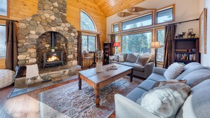A cozy living room with a stone fireplace, wooden ceiling, large windows, and comfortable seating. - Forest Views, Hot Tub & Tahoe Donner Access | Alpenglow Chalet by AvantStay (Truckee)