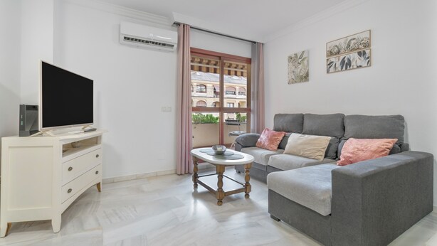 A living room with a grey sofa, a white TV stand, a small round table, and a window with a view of buildings.