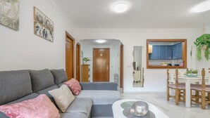 A living room with a grey sofa, a dining table with chairs, a kitchen area with cabinets, and a hallway leading to a door.