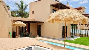 Un área de piscina moderna con un jacuzzi, mobiliario exterior y una sombrilla de paja.