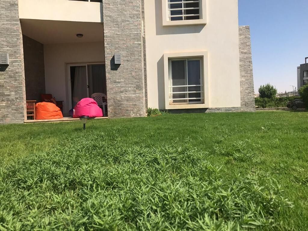A modern building with a flat roof, a balcony with orange bean bag chairs, and a well-maintained lawn.