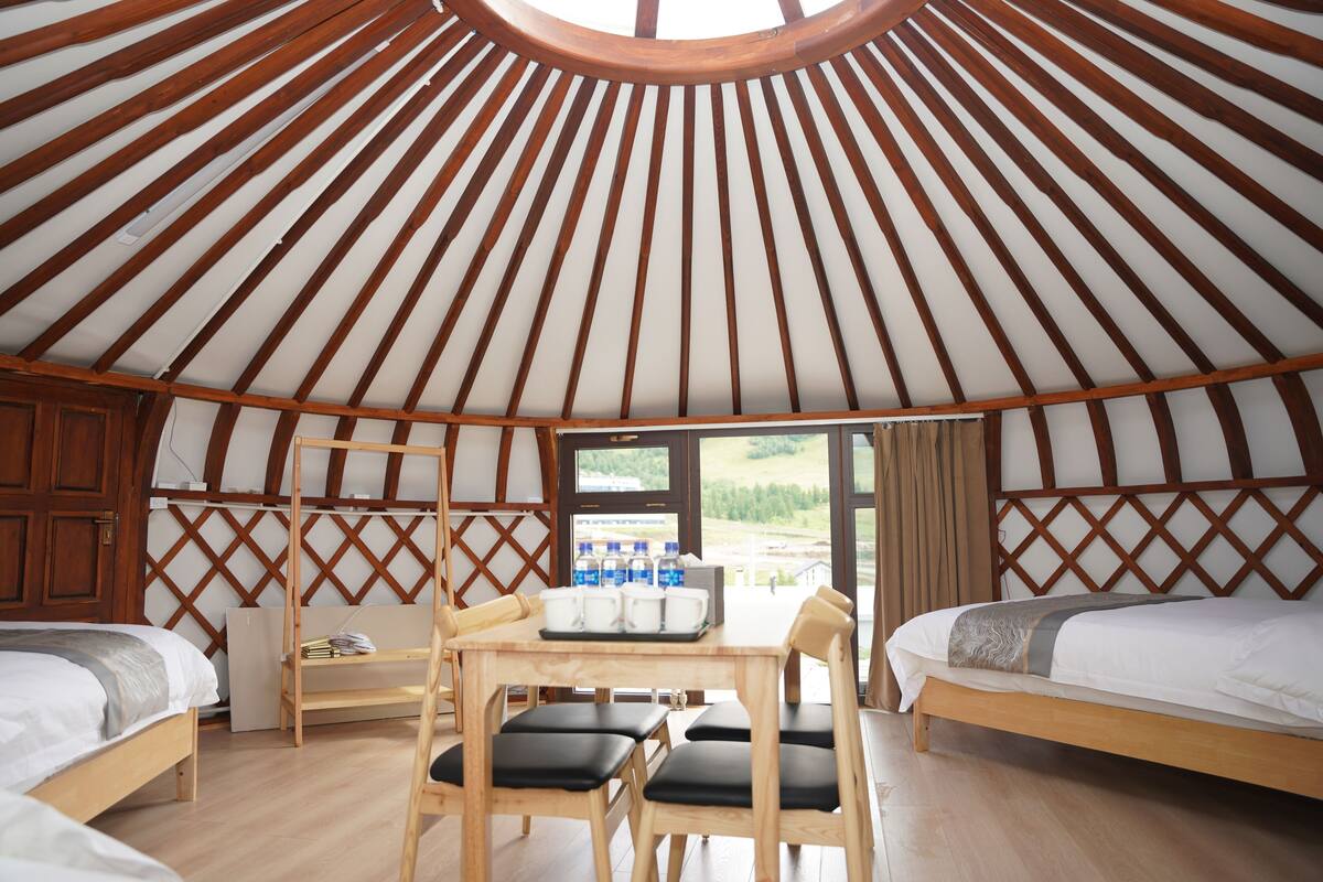 Een joert met twee bedden, een houten tafel met stoelen, en een raam met uitzicht op de omgeving.