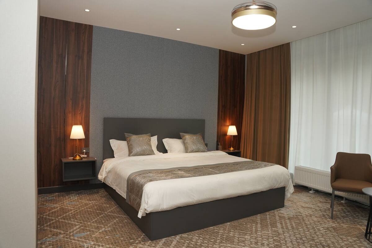 Een moderne hotelkamer met een groot bed, twee nachtkastjes met lampen, een stoel en een raam met gordijnen.