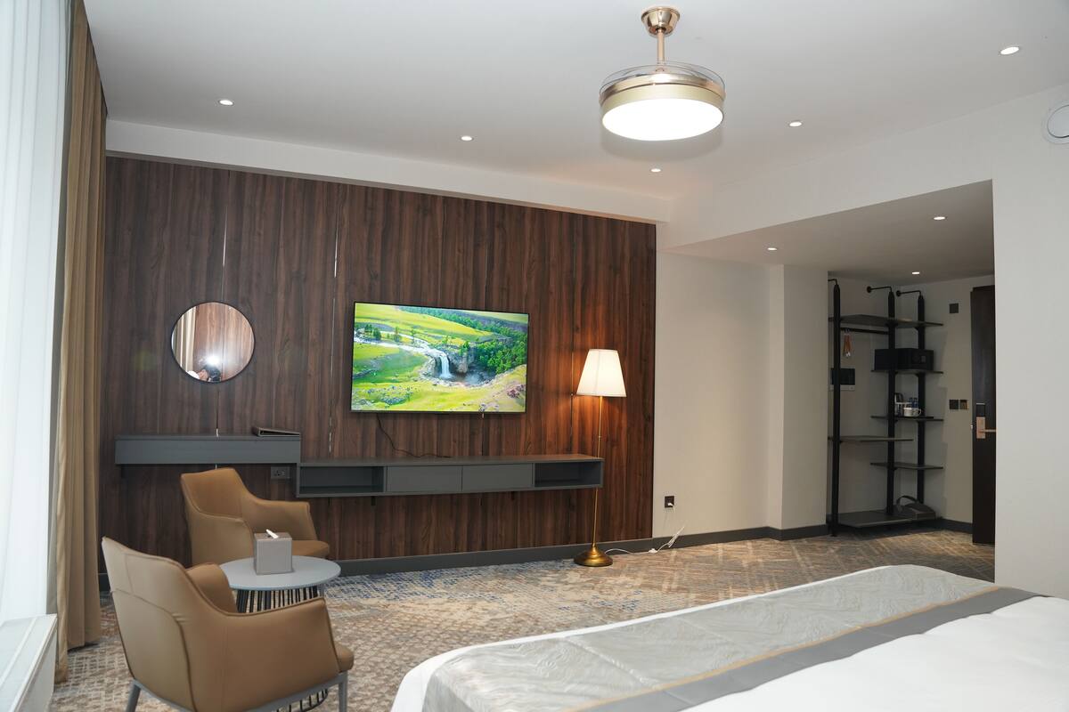 Een moderne hotelkamer met een groot bed, een televisie bevestigd aan een houten wand, een bureau met een lamp en een zithoek met stoelen.