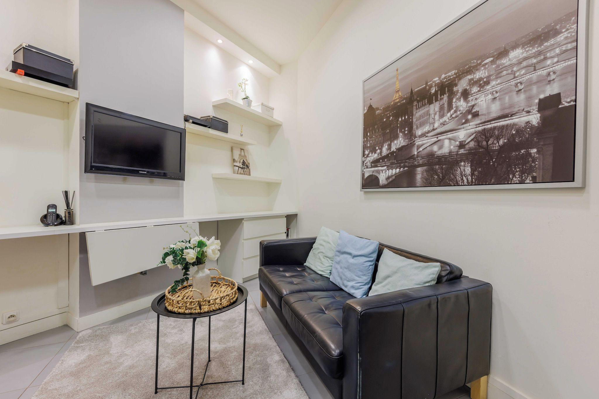 A modern living room with a black leather sofa, a flat-screen TV mounted on a white wall, and a large black-and-white cityscape painting.