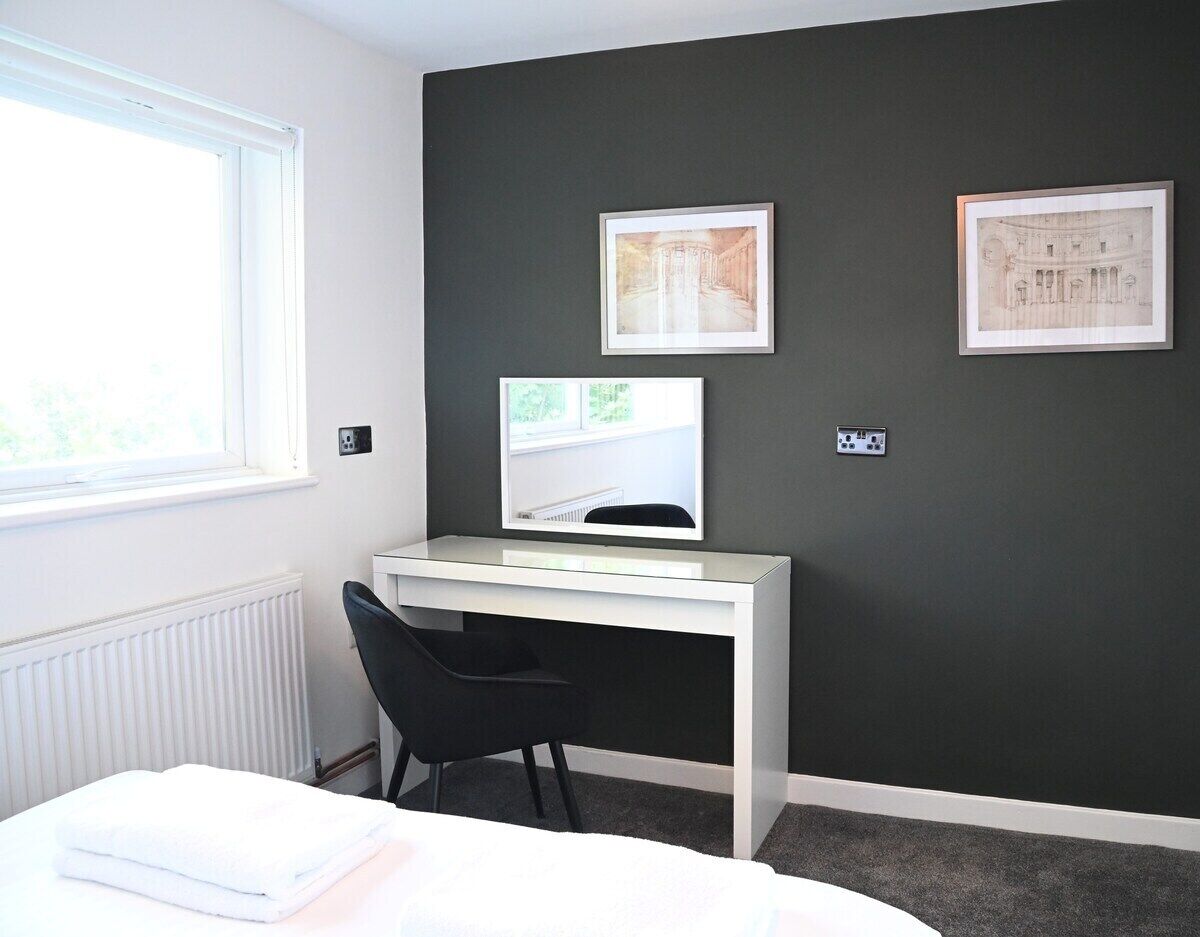 Habitación de hotel moderna con escritorio blanco, silla negra y cama con edredón blanco.