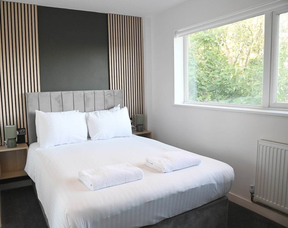 A neatly made bed with white linens and towels, a headboard with vertical slats, bedside tables with lamps, and a window with a view of greenery.