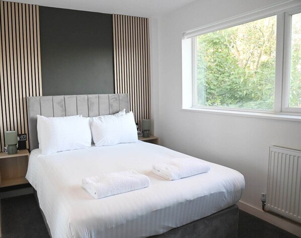 Una cama bien hecha con sábanas y toallas blancas, cabecera con listones verticales, mesitas de noche con lámparas y una ventana con vistas a zonas verdes.