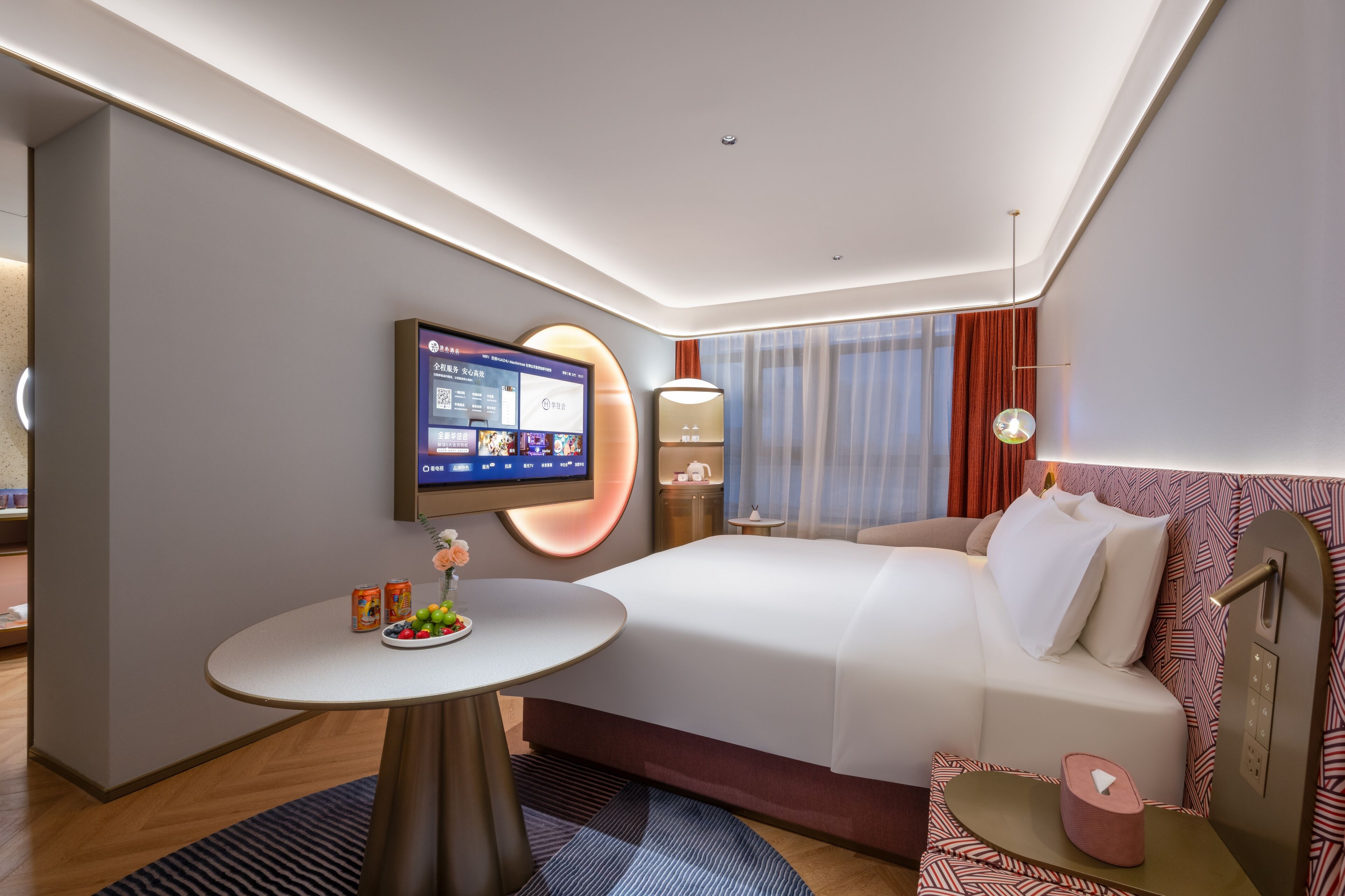 A modern hotel room with a large bed, a flat-screen TV mounted on the wall, a small round table with snacks, and a bedside table with a lamp.