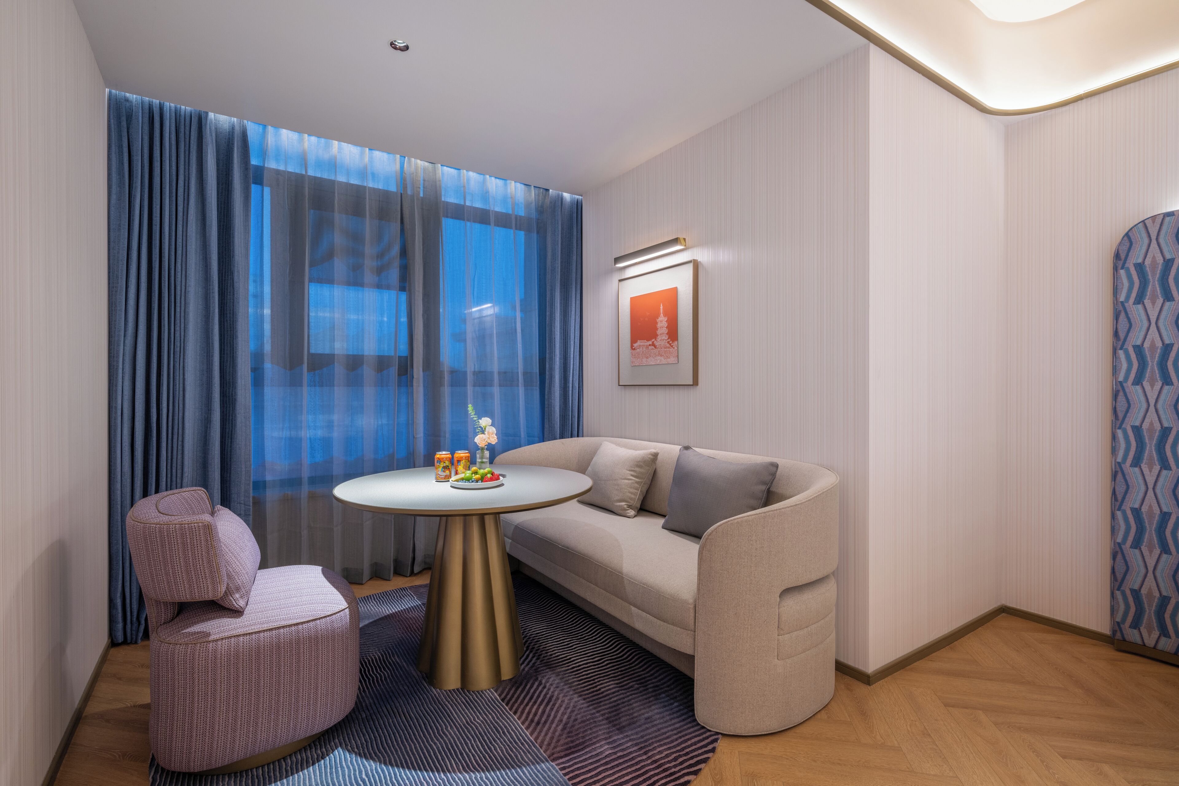 A modern hotel room with a sofa, a small round table with a vase and fruit, a pink chair, and a large window with blue curtains.