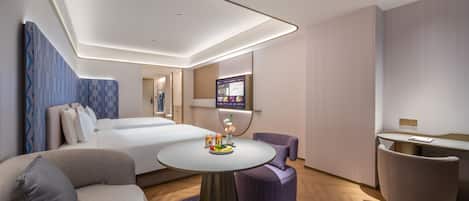 Ein modernes Hotelzimmer mit einem Bett, einem Sofa, einem runden Tisch mit Obst, einem Fernseher und einer kleinen Sitzecke.