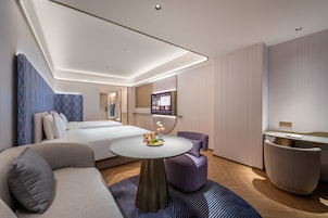 Quarto de hotel moderno com cama, sofá, mesa redonda com frutas, televisão e um pequeno espaço para sentar.