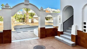 Un espacio exterior con piso de baldosas blancas y marrones, una escalera con barandilla blanca y vistas a palmeras y edificios al fondo.