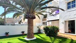 Un patio cuidado con una palmera, césped artificial, plantas en macetas y un edificio blanco con balcones.