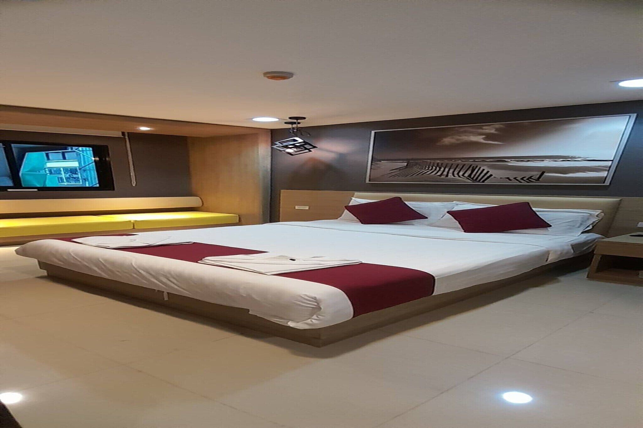 Una cama grande con ropa de cama roja y blanca, un televisor de pantalla plana a la izquierda y una obra de arte colgada en la pared sobre la cama.