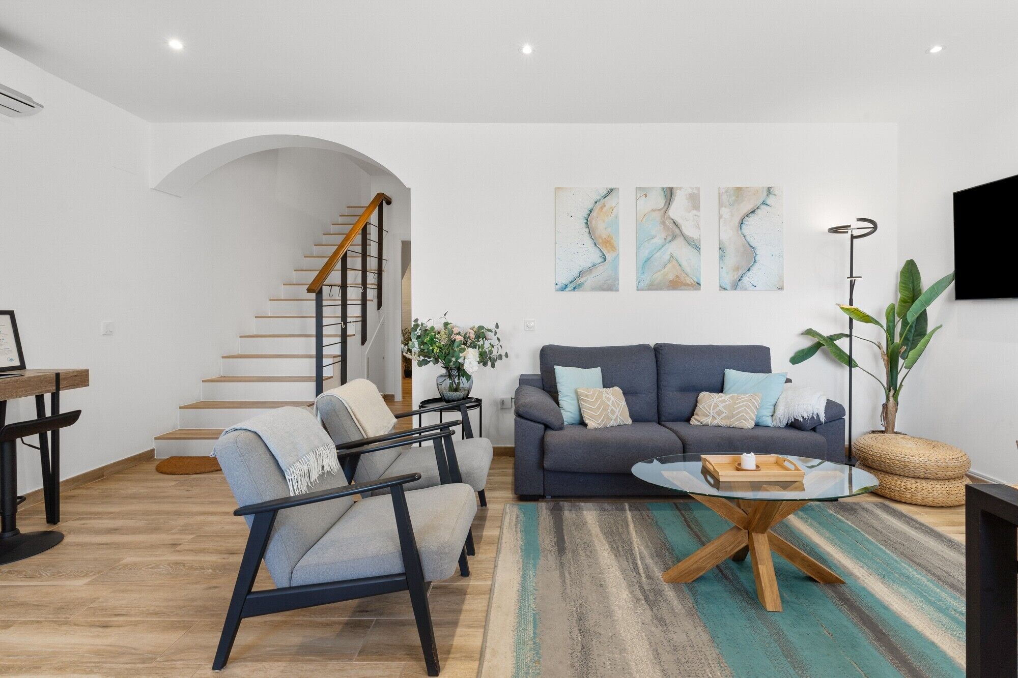 Una sala de estar moderna con una escalera, un sofá gris, una mesa de centro redonda y una planta grande.