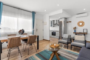Ein modernes Wohnzimmer mit einem Glastisch, Korbstühlen, einer Couch und einer Küche im Hintergrund.