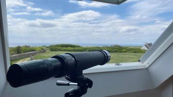 A telescope on a tripod in a room with a skylight, overlooking a green landscape and a cloudy sky.
