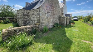 Un bâtiment en pierre doté d’un toit en pente, entouré d’une zone herbeuse agrémentée de fleurs et d’un mur de pierre.