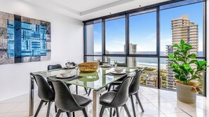Un espace repas moderne avec une table en verre, des chaises noires et une vue sur l’océan et les bâtiments.