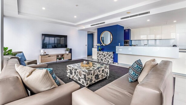 Un salon moderne doté d’un grand téléviseur à écran plat, de sièges confortables, d’une table basse à motifs et d’un coin cuisine avec des meubles blancs et des touches de couleur bleue.