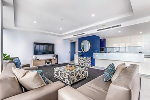 A modern living room with a large flat-screen TV, comfortable seating, a patterned coffee table, and a kitchen area with white cabinets and blue accents. - Aria 1903 (Broadbeach)