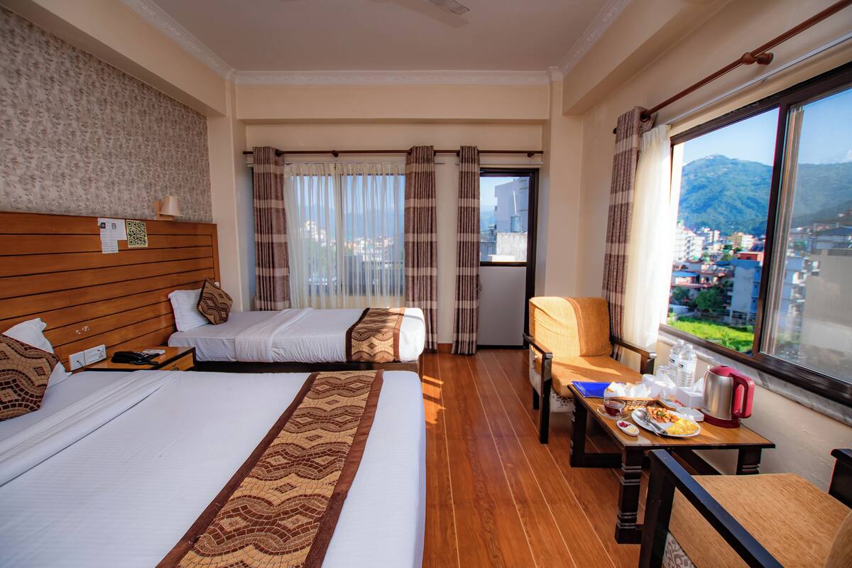 A hotel room with two beds, a dining table with breakfast, a view of the city and mountains, and a window with curtains.