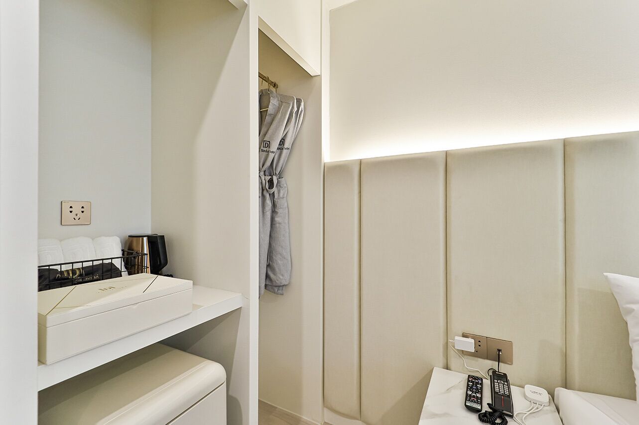 A hotel room with a white vanity, a robe hanging on the door, a bed with a white pillow, and a bedside table with a phone and a charger.