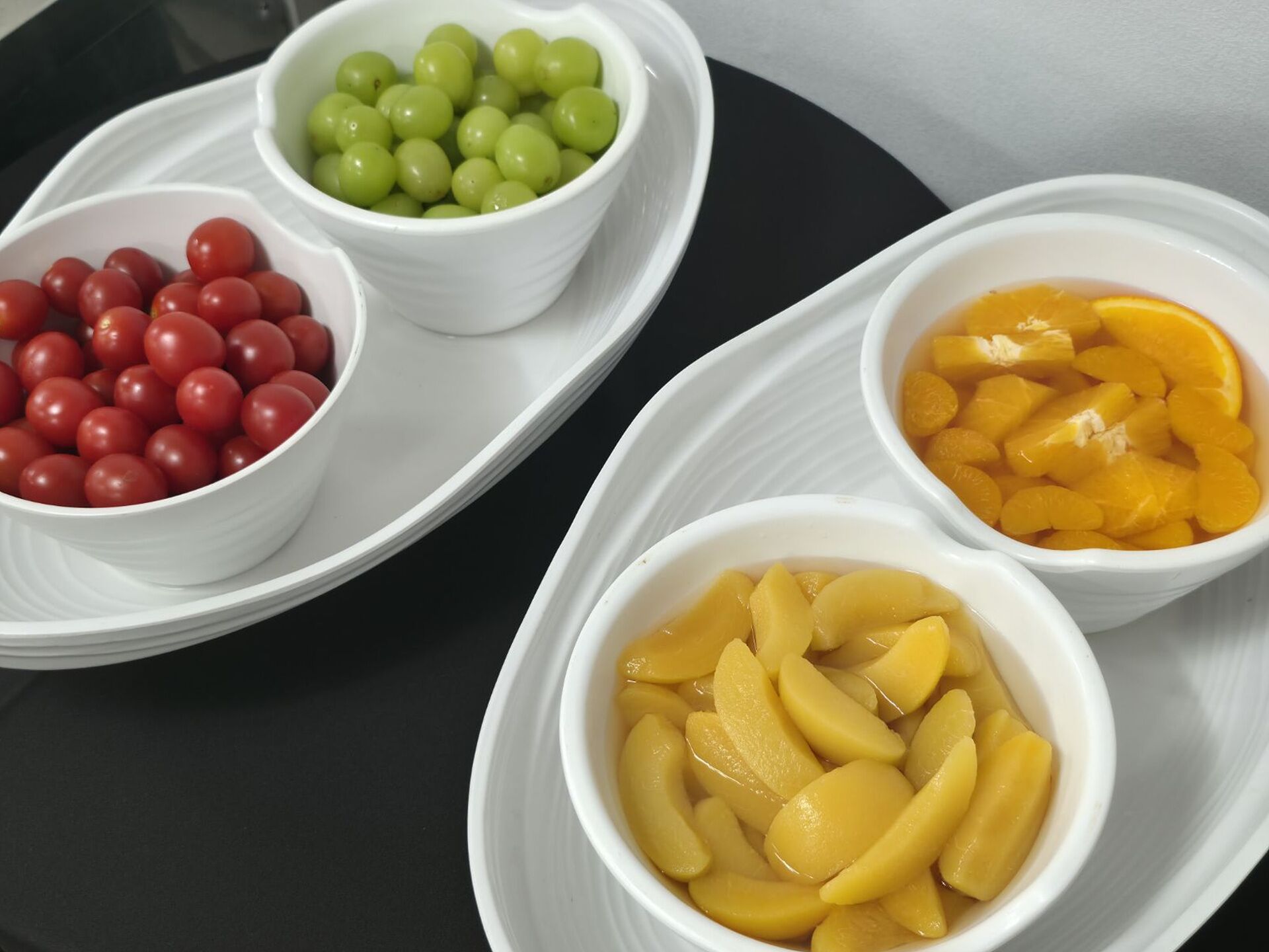 Three white bowls on a black tray containing grapes, cherry tomatoes, and sliced peaches.