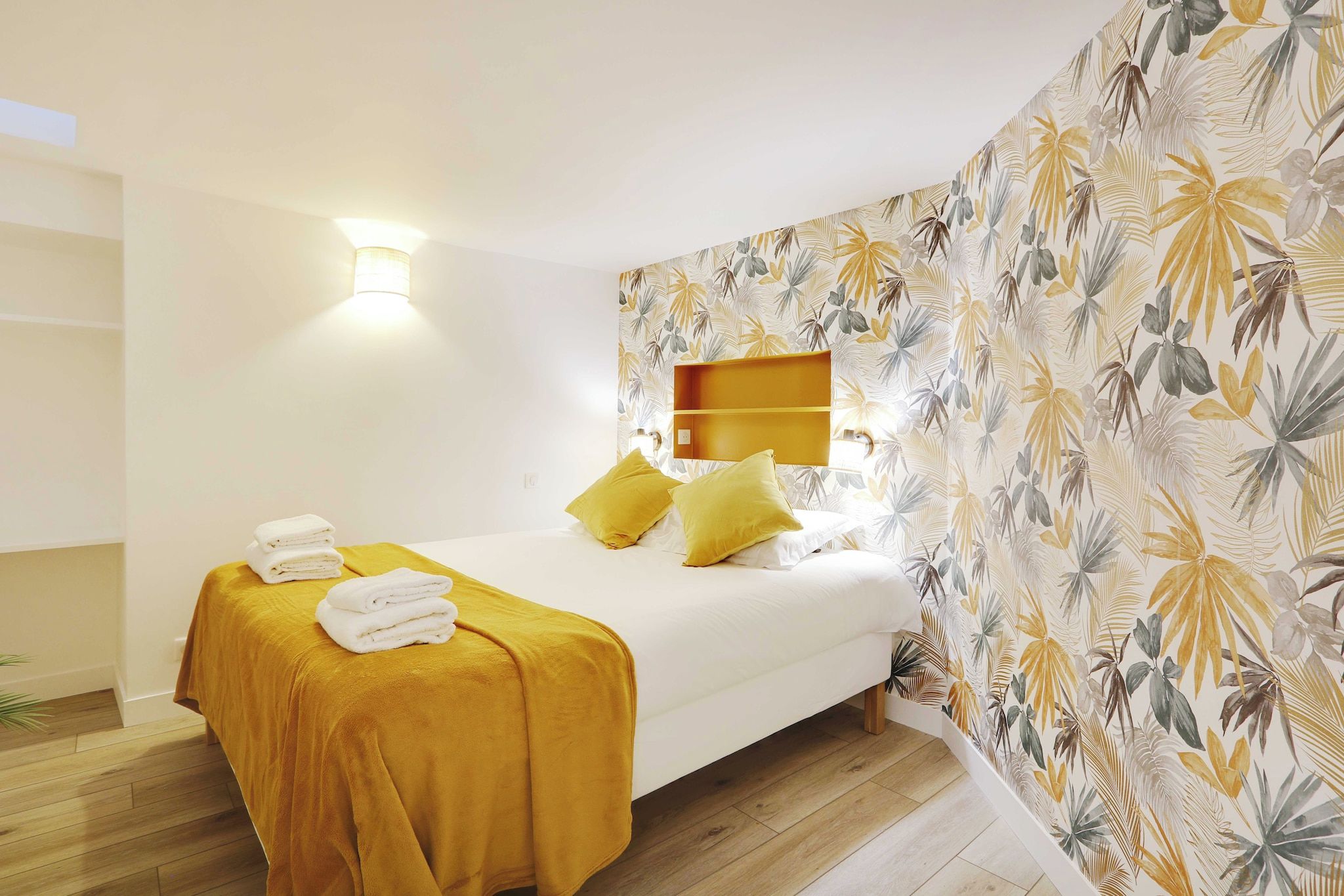 Une chambre d’hôtel avec un lit, une table de chevet, une étagère fixée au mur et un papier peint à motifs.