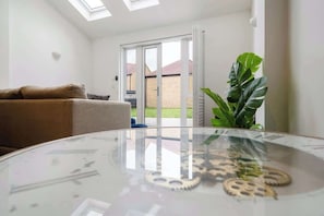 A modern living room with a glass table reflecting ceiling lights, a brown sofa, and a large green plant.