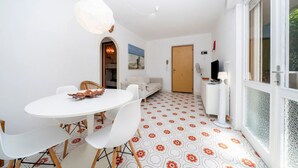 A dining area with a white table and chairs, a hanging light fixture, a patterned floor, a sofa, and a door in the background. - GuestHost - Cozy 56 sqm ground-floor apartment featuring a balcony running along the entire perimeter and a private garden, perfect for relaxation and tranquility. Located in Grado Pineta, a green and quiet area just steps from the sea and nature trails, (Grado-Pineta)