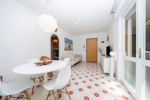 Un área de comedor con una mesa y sillas blancas, una lámpara colgante, piso estampado, un sofá y una puerta al fondo.