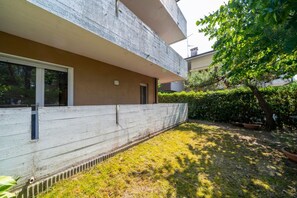 Un edificio moderno con fachada de concreto, un ventanal pequeño y un patio ajardinado con un árbol.