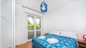 A bedroom with a bed, a door, a window, and a chandelier. - GuestHost - Cozy 56 sqm ground-floor apartment featuring a balcony running along the entire perimeter and a private garden, perfect for relaxation and tranquility. Located in Grado Pineta, a green and quiet area just steps from the sea and nature trails, (Grado-Pineta)