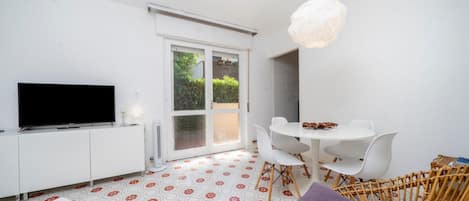 A modern living room with a flat-screen TV, white furniture, a dining table, and a wicker chair.