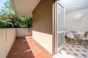 Un balcón con suelo de baldosas, una puerta de cristal que da a una habitación con una mesa y sillas blancas, y vistas a árboles.