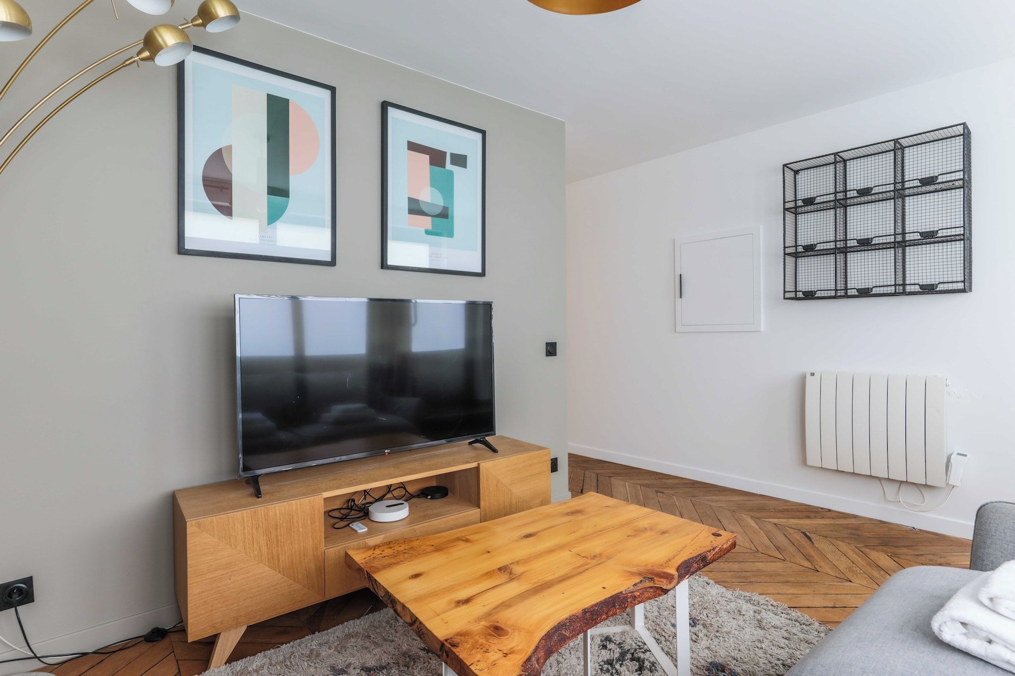 A modern living room with a flat-screen TV on a wooden console, two abstract paintings on the wall, a black metal rack, and a radiator.